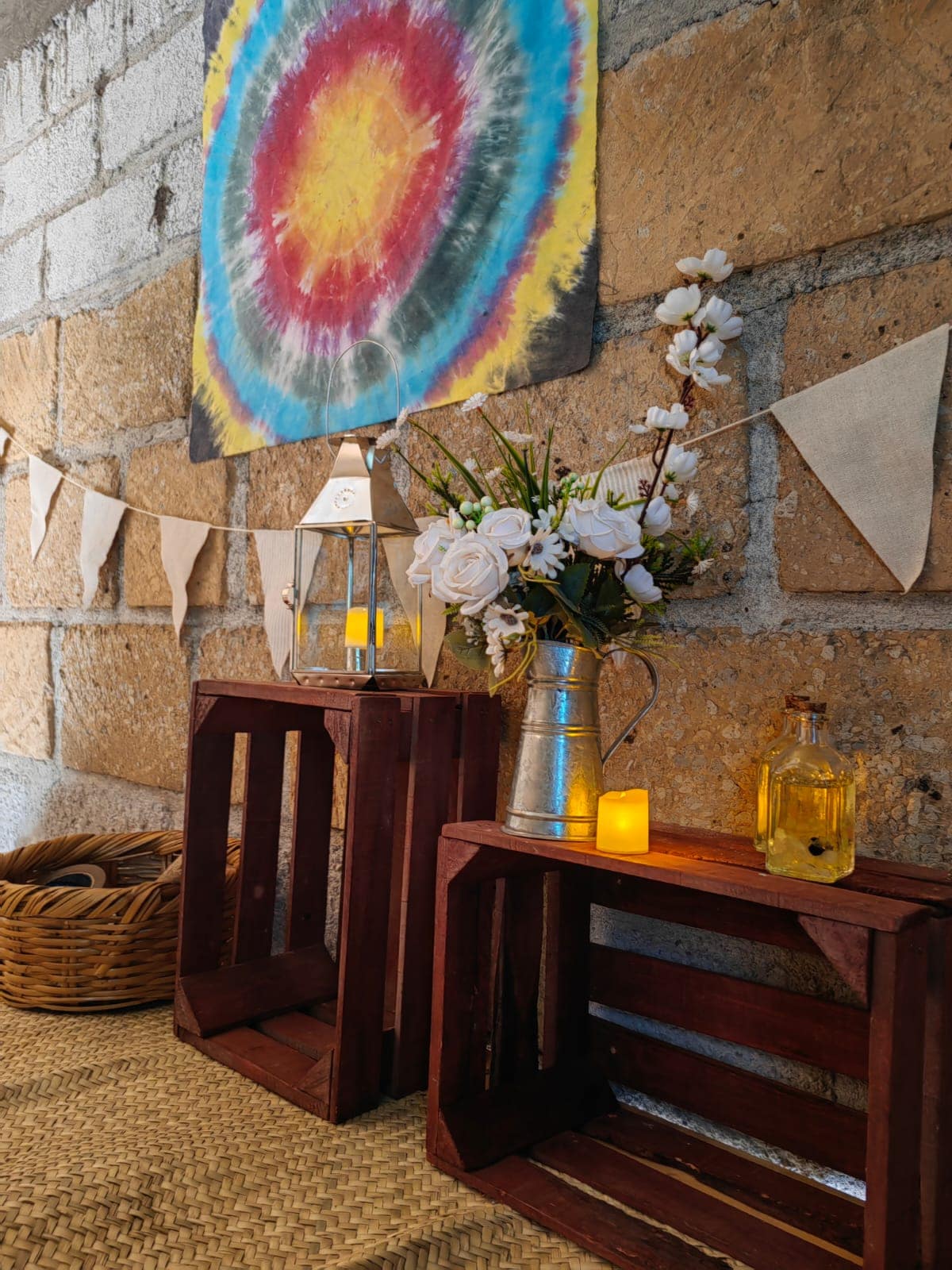 A rustic display features two wooden crates with a silver pitcher filled with white flowers and accompanied by decorative glass bottles. A lantern rests on one crate, alongside soft lighting from candles. Colorful wall art and fabric bunting enhance the warm and inviting space.