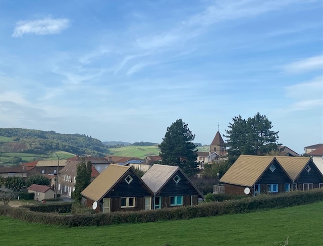 Chalets with sloped roofs are positioned in a lush green landscape, surrounded by gentle hills. In the background, a village with a church steeple is visible among trees, under a clear blue sky.