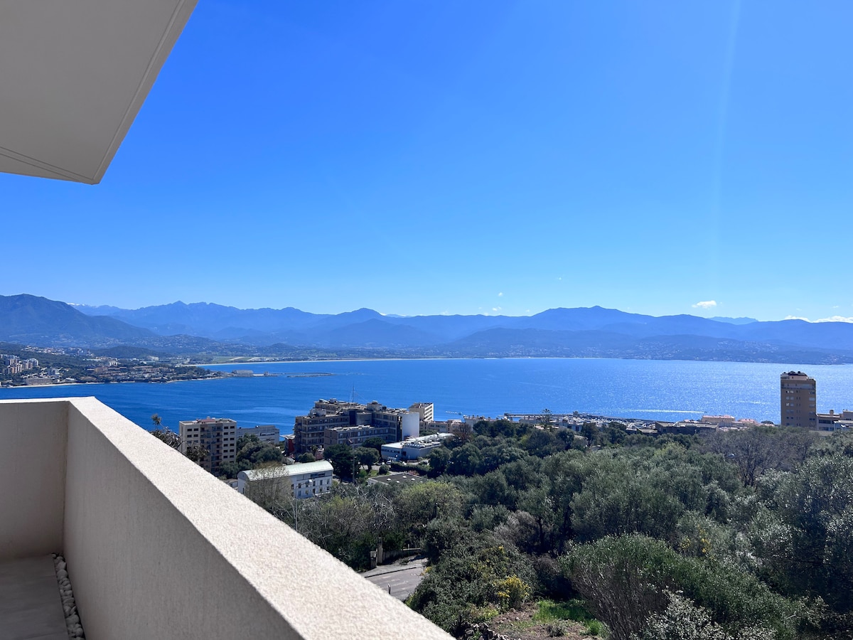A panoramic view of the coastline is captured from a spacious terrace. Clear blue skies and distant mountains frame the shimmering blue sea, while the landscape below features a mix of greenery and urban structures.