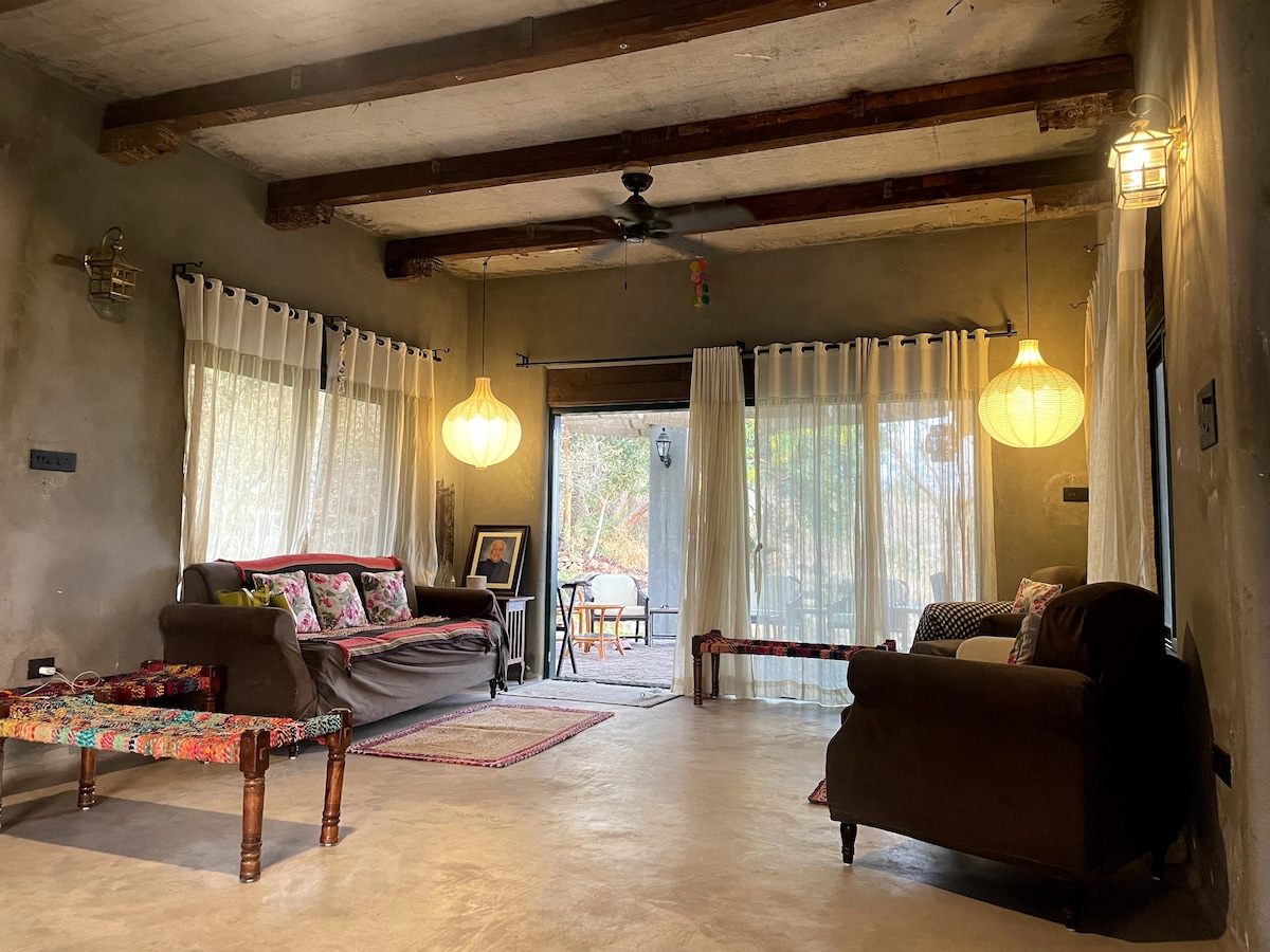 A cozy living area features two comfortable sofas, draped with decorative pillows, complemented by a colorful tapestry. Large windows allow natural light to fill the space, while soft lanterns provide a warm glow. A wooden table is visible through the open doorway, surrounded by greenery.