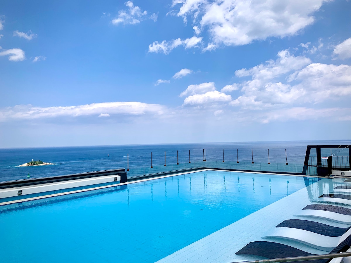 An infinity pool extends towards the horizon, offering panoramic views of the ocean and sky. The calm waters reflect the clouds above, creating a serene atmosphere. Lounge chairs line the pool's edge, inviting relaxation in this scenic setting.
