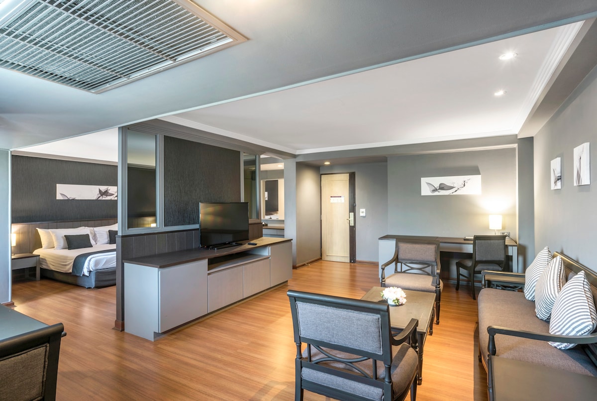 A spacious living area features a modern entertainment unit with a flat screen TV. Comfortable seating is arranged around a coffee table, with artwork displayed on the gray walls. The bedroom area is separated, showcasing a king-sized bed and additional lighting.