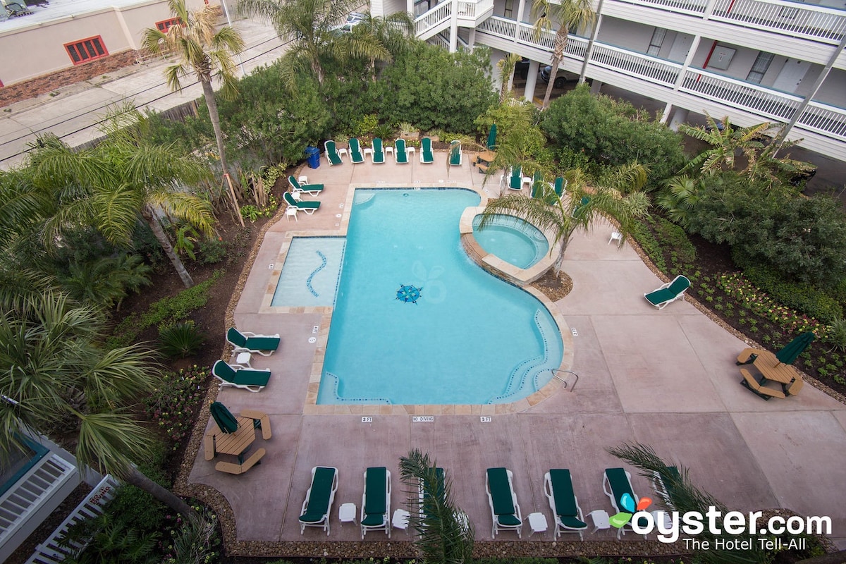 The outdoor pool area is surrounded by lush greenery and features a main pool shaped like a figure eight, along with a separate hot tub. Multiple lounge chairs are positioned around the pool deck, inviting relaxation under the sun.