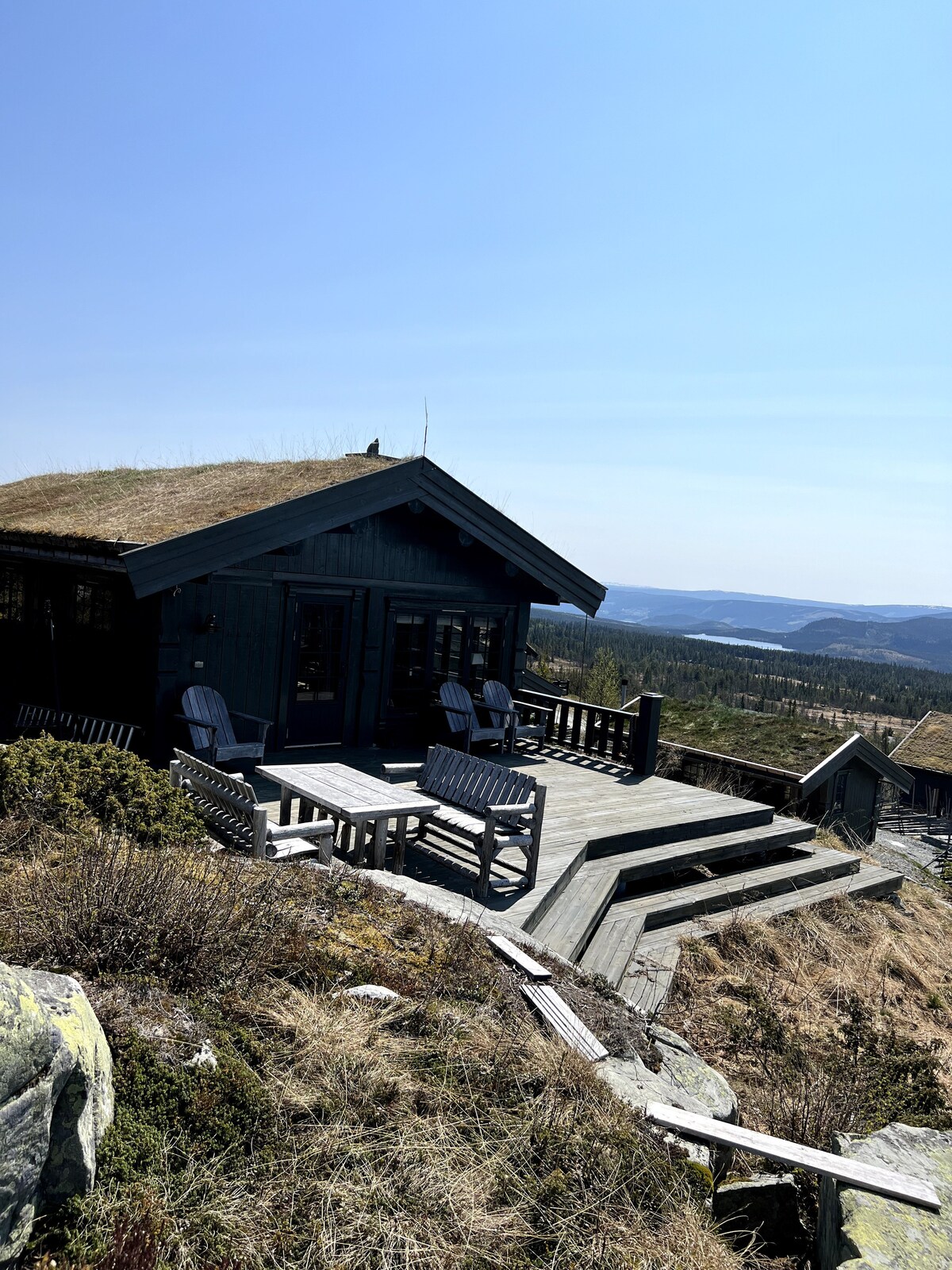 A modern cabin is set against a mountainous backdrop, featuring a wooden deck with several outdoor chairs. The sloped roof is covered with grass, adding to the natural aesthetic. Expansive views of the surrounding landscape are visible in the distance.