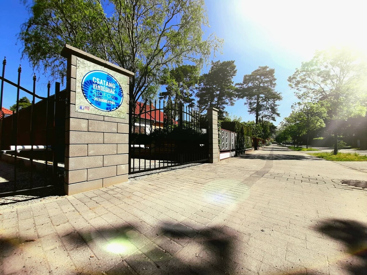 An entrance gate is featured, surrounded by a stone wall. The gate opens onto a tree-lined pathway, with sunlight filtering through the leaves. A blue signplate with clear text indicates the property's name. The atmosphere conveys a peaceful neighborhood setting.