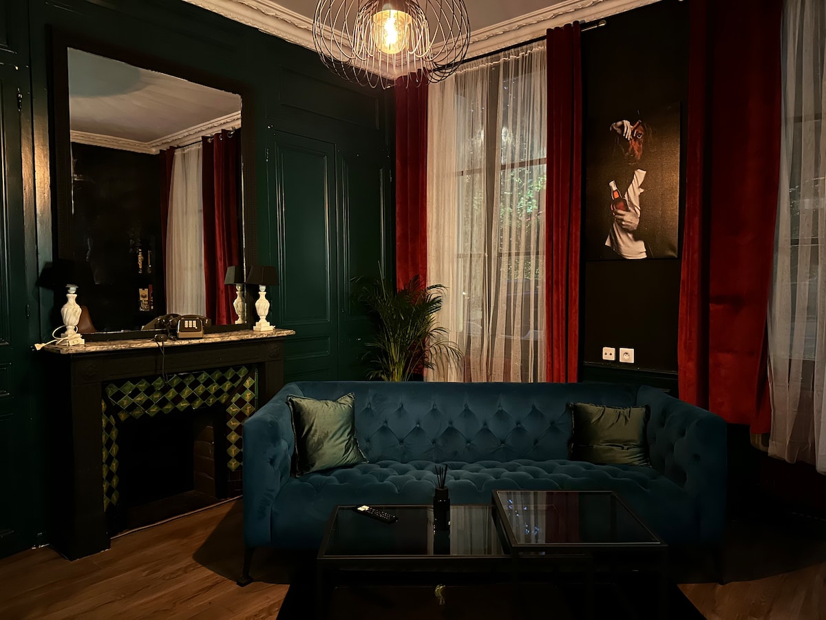 A stylish living area features a deep blue velvet sofa with decorative cushions, paired with a modern coffee table. Rich green walls and deep red curtains create a bold contrast, while a detailed artwork and a fireplace are visible, adding character to the space.