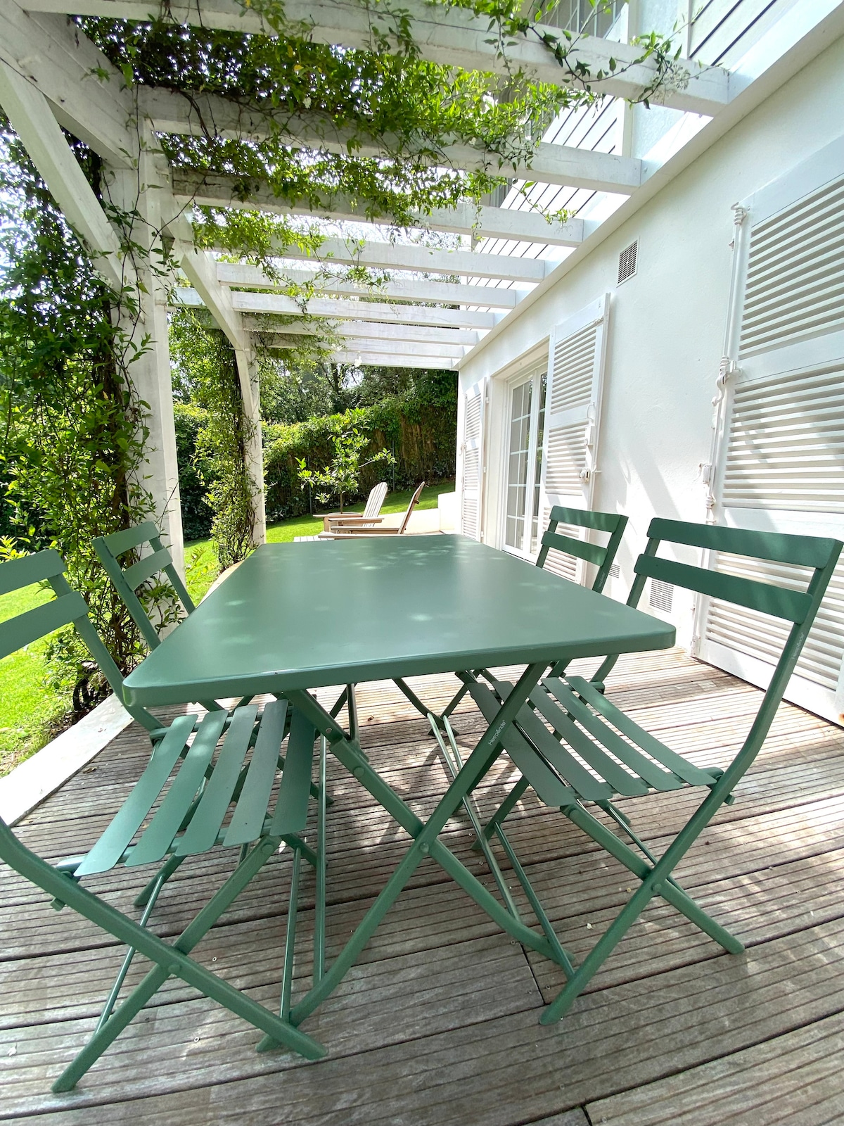 An outdoor terrace features a green metal table surrounded by four matching folding chairs. Sunlight filters through a white trellis, while lush greenery and a lawn are visible in the background. Light-colored shutters are present on the exterior wall.