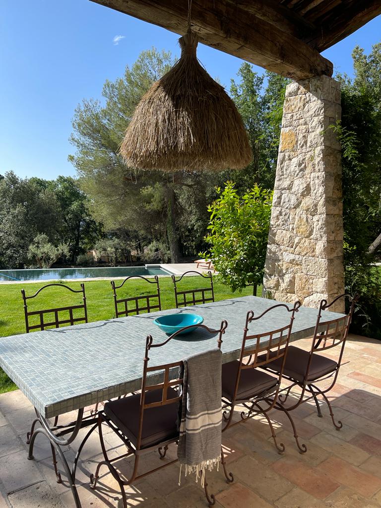 A dining table with metal chairs sits beneath an overhead straw fixture, surrounded by greenery. The view includes a grassy area leading to a serene pool in the background, offering a tranquil outdoor dining space.
