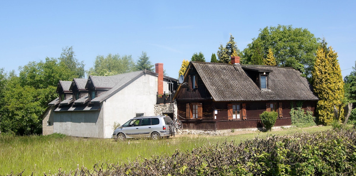 A spacious property is set among lush greenery, featuring a combination of modern and traditional architecture. The wooden structure showcases a thatched roof, while the adjacent gray building presents a contemporary design. A vehicle is parked in front, with a hedge delineating the area.