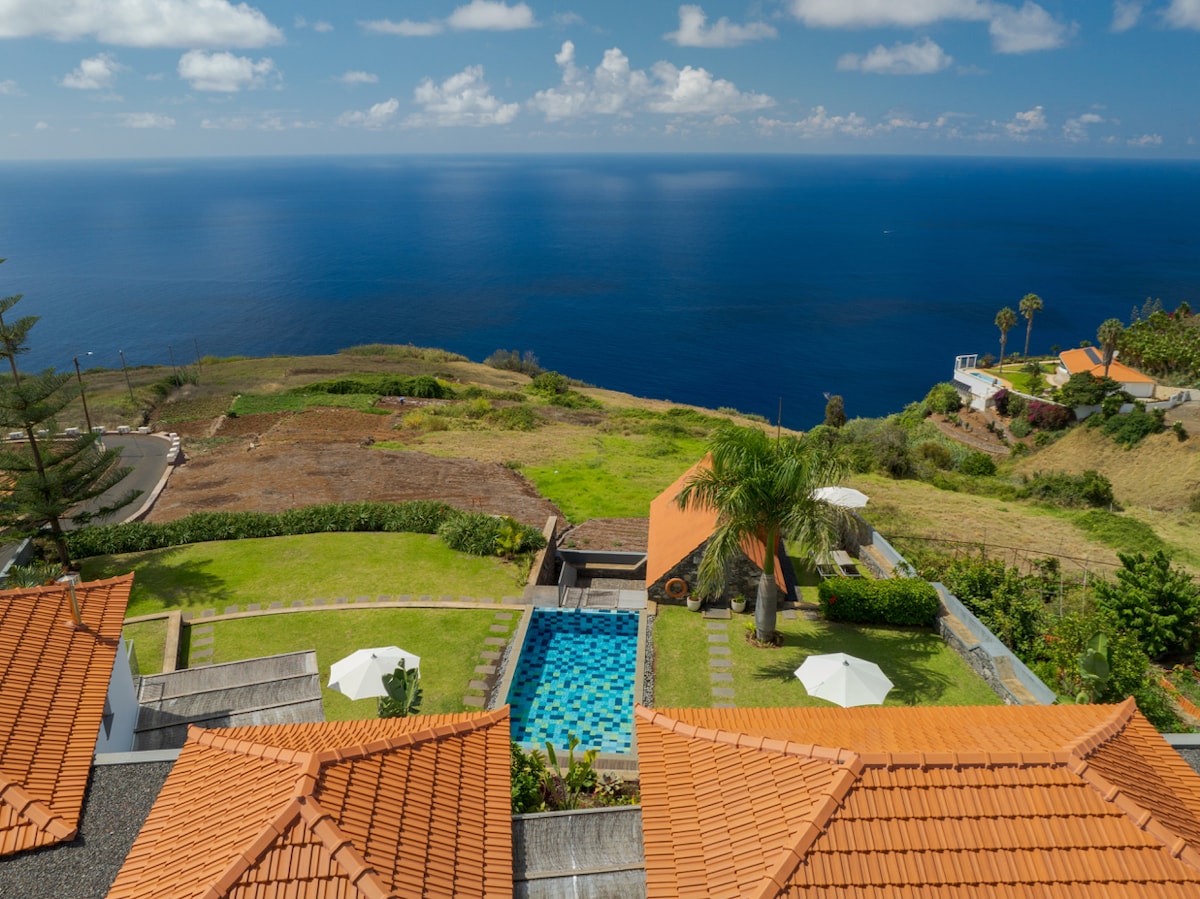 An aerial view captures the heated swimming pool nestled in a lush garden. The vibrant blue of the pool contrasts with the expansive Atlantic Ocean and the scenic landscape. Two sun loungers and sun umbrellas are positioned around the pool, enhancing the outdoor relaxation space.