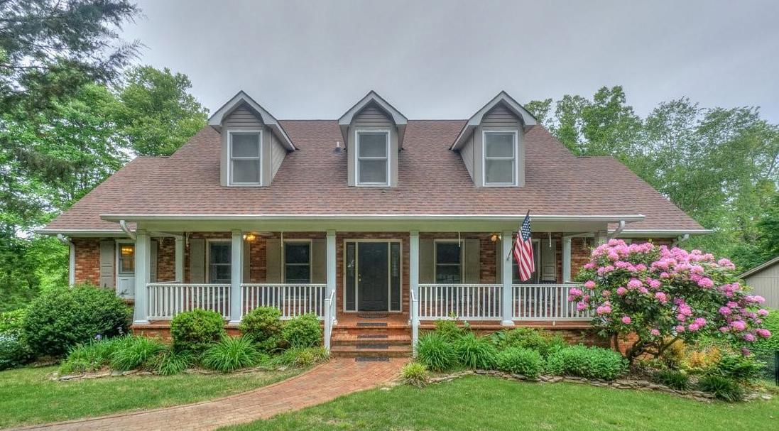 A two-story house with a brown shingle roof and symmetrical gabled windows is framed by green landscaping. A covered porch, with white railings, invites relaxation. A flag is displayed to the right, and blooming bushes add a touch of color alongside the path leading to the entrance.
