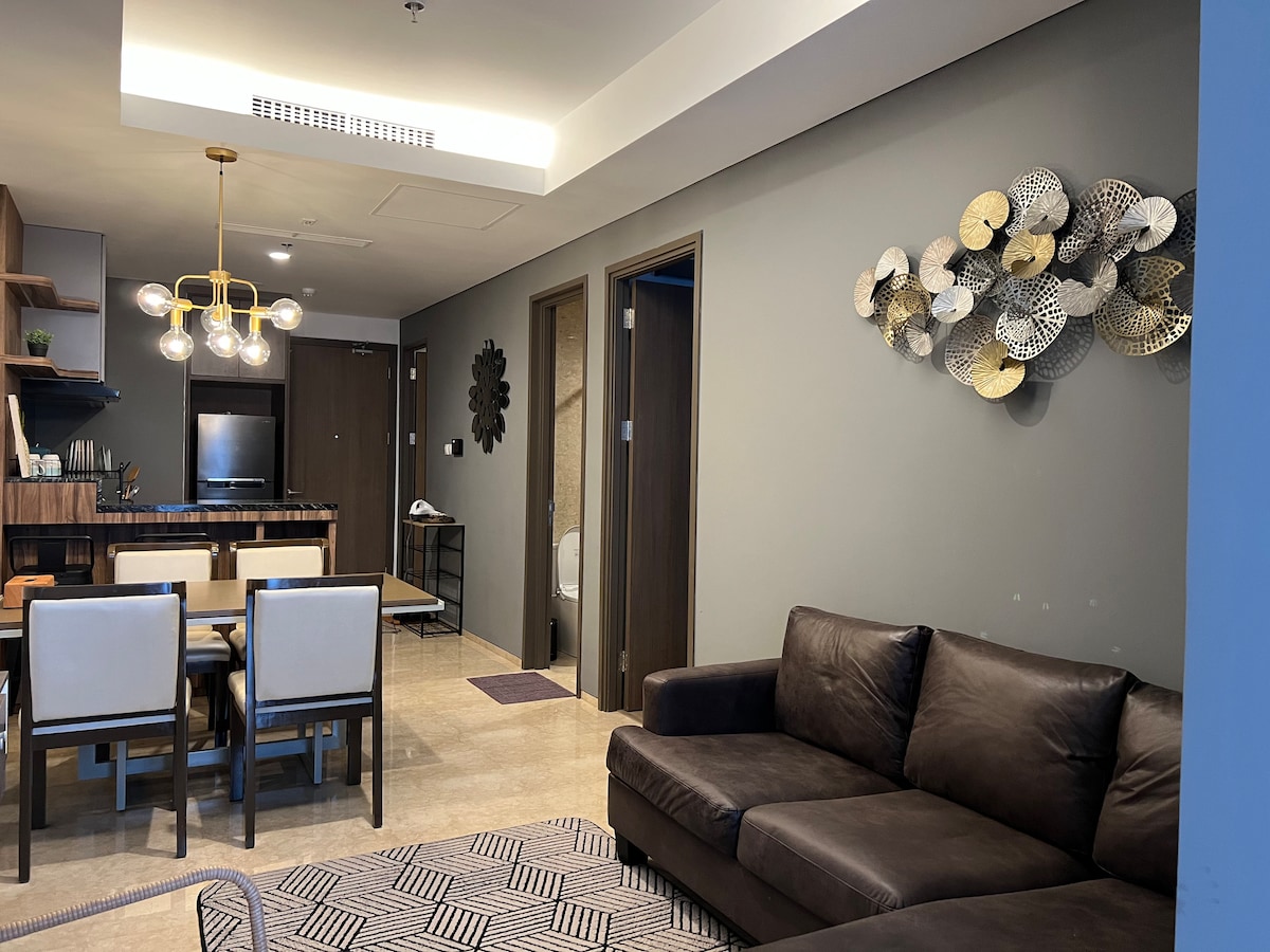 A modern living area features a dark leather sectional sofa and a dining table surrounded by four light-colored chairs. Decorative wall art made of metallic discs adds visual interest to the space, while a flat-screen television is visible in the background.