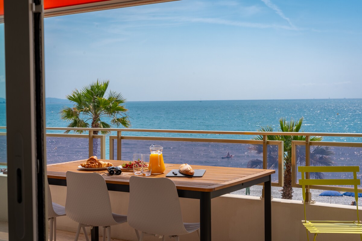 A dining table is positioned on a balcony, offering a view of the sea. Brightly colored palm trees line the beach, while the table is set with a pitcher of orange juice, fresh pastries, and a bowl of fruit. The sky is clear with soft clouds.