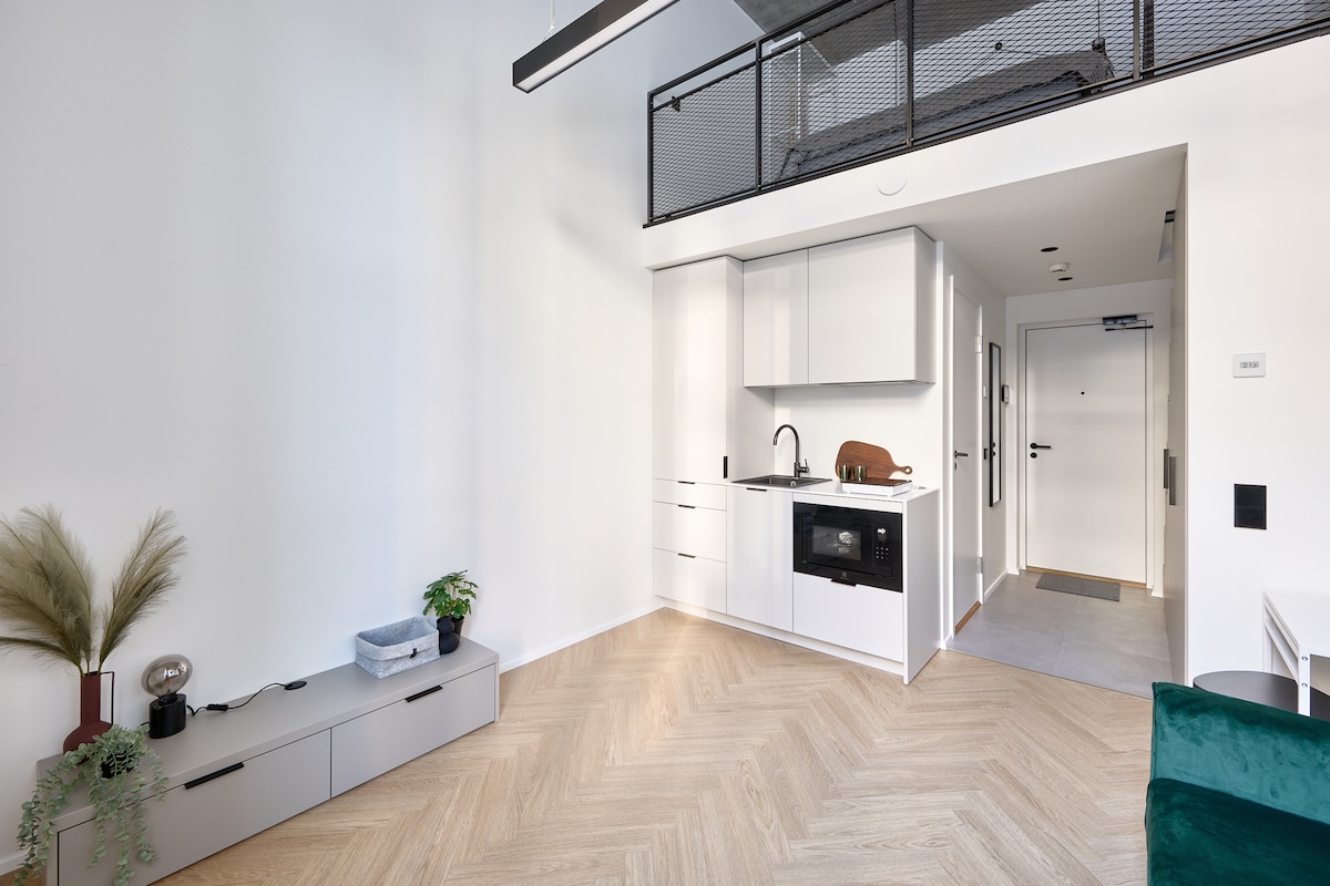 A well-lit space showcases a modern kitchenette with sleek cabinetry and integrated appliances. Light wood flooring complements the airy atmosphere. A small dining area is visible, alongside a decorative shelf with potted plants, enhancing the minimalist design.