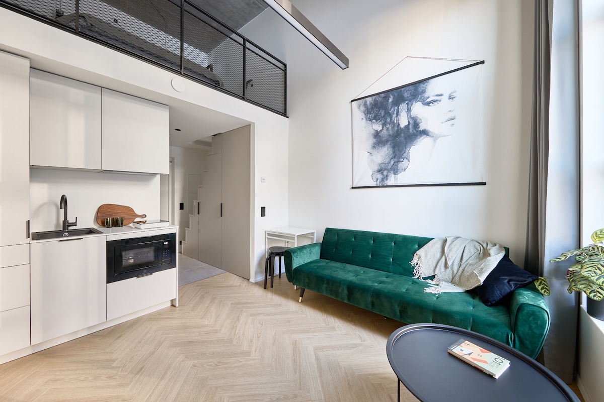 A well-designed kitchenette features modern appliances, including a microwave and a stovetop. A green velvet sofa adds a touch of elegance, paired with a black side table. A piece of abstract wall art enhances the space, while natural light filters through large windows.