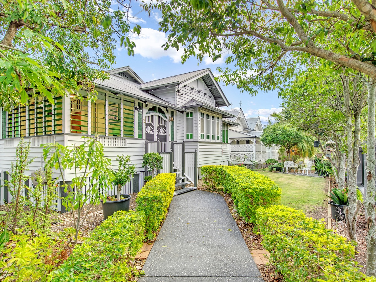 A charming entryway is framed by lush greenery and low hedges, leading to the inviting façade of the historic cottage. The exterior features distinctive architectural details, large windows with decorative shutters, and a welcoming pathway that guides guests toward the front door.