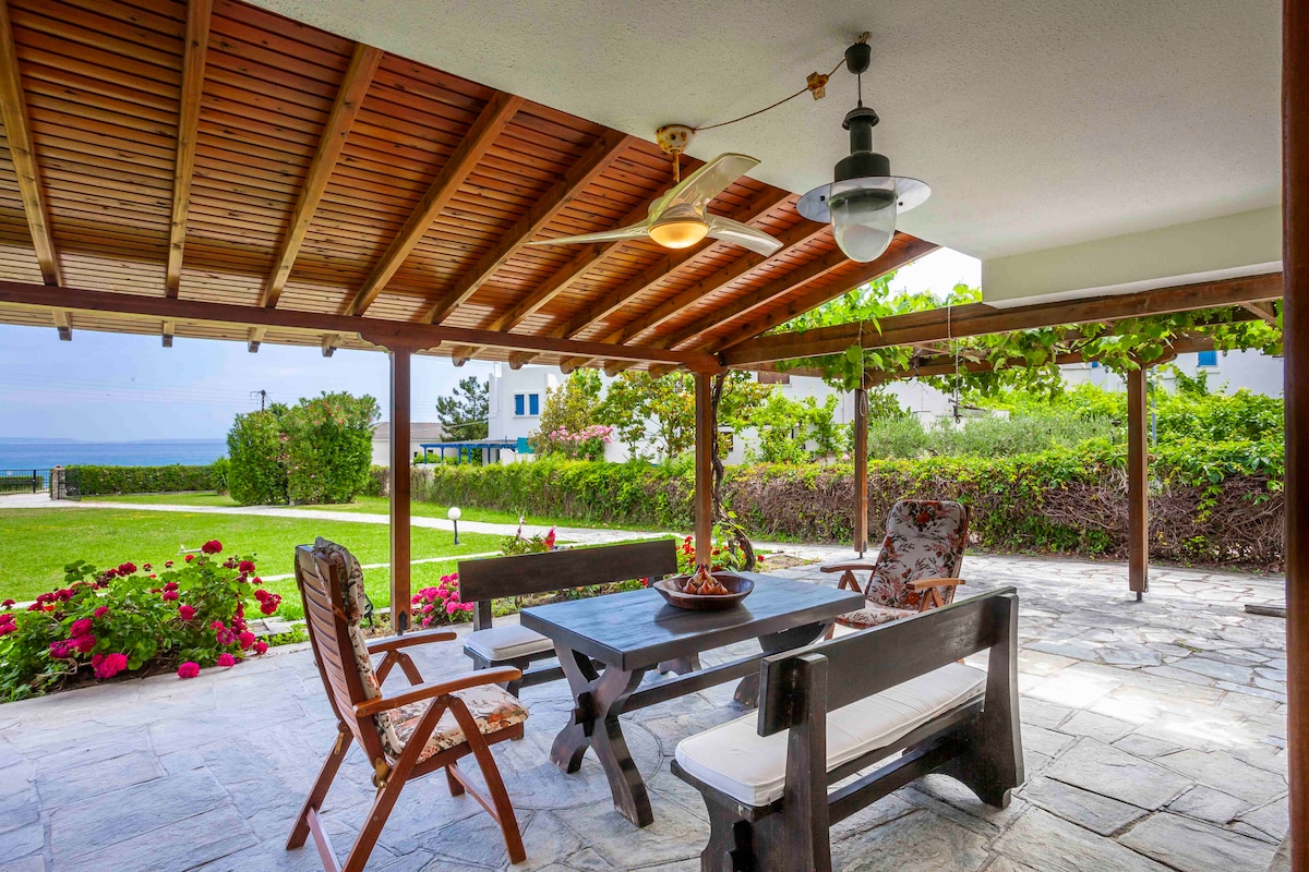 An outdoor dining area covered by a wooden pergola features a large dark wooden table surrounded by cushioned chairs. Colorful flowers are visible in the surrounding garden, and the sea can be glimpsed in the distance through the greenery.