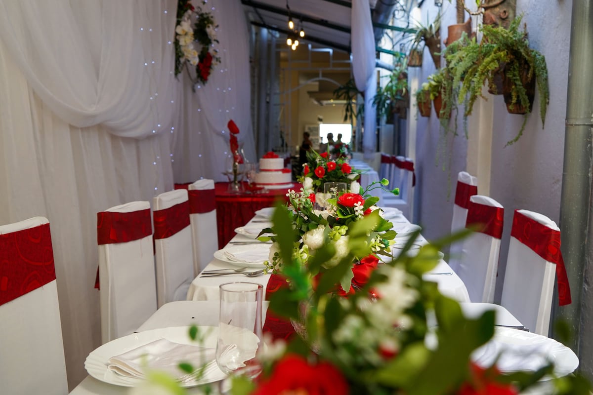 A long dining table is set with white tablecloths and red accents, featuring floral arrangements and elegant dinnerware. Soft lighting illuminates the pathway, where greenery is hung, creating a vibrant and welcoming dining environment suitable for both traditional and international cuisine.