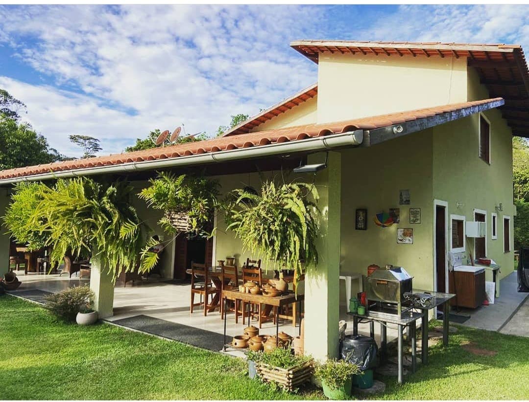 A spacious outdoor area is featured, surrounded by vibrant greenery and a well-maintained lawn. The building showcases a terracotta roof and large windows, while wooden dining furniture is arranged under an awning. Potted plants and decorative items enhance the inviting space.