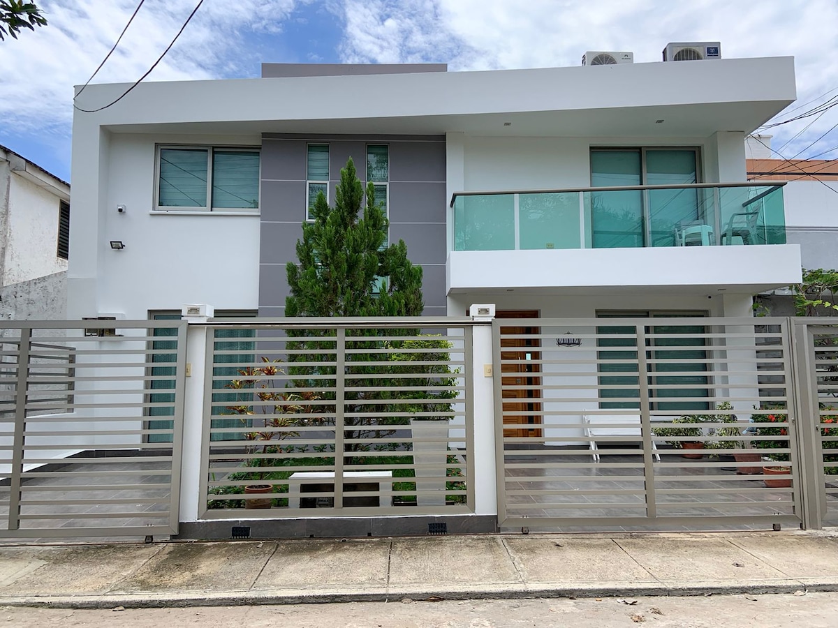 The modern exterior of the house is highlighted by clean lines and a sleek design. A private entrance is visible through a decorative gate, flanked by planted greenery and seating areas. Large windows facilitate natural light, while a balcony adds a touch of spaciousness to the facade.