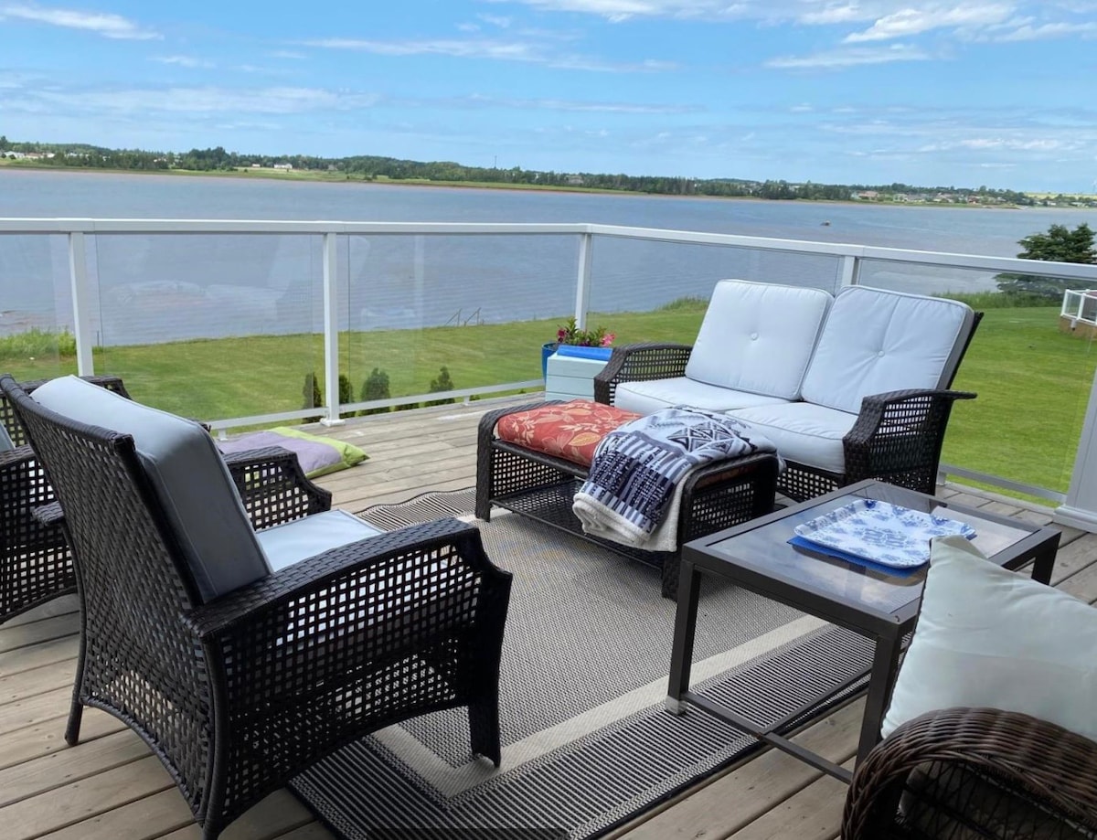 A spacious deck features comfortable seating arrangements, including two armchairs and a loveseat, overlooking the tranquil waters of the bay. A coffee table is placed centrally, with a patterned throw draped over the loveseat, inviting relaxation while enjoying scenic views.