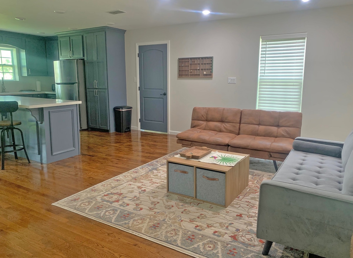 A spacious living area is presented, featuring a brown leather couch and a gray sofa, positioned around a coffee table. Natural light enters through a window adorned with light blinds. The open layout connects to a well-equipped kitchen, enhancing the home's inviting ambiance.