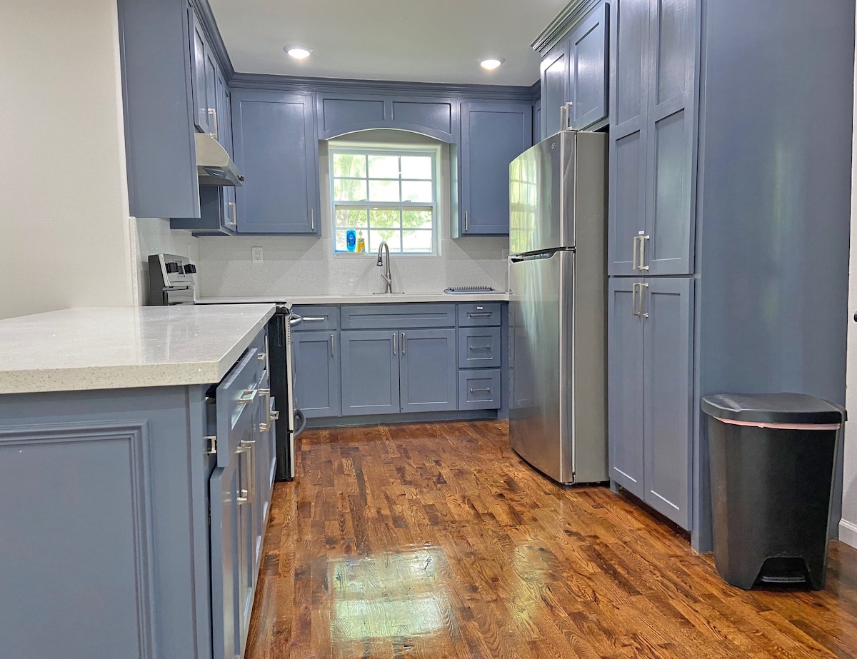A modern kitchen features blue cabinetry and sleek stainless steel appliances. A large window allows natural light to brighten the space, while a polished countertop provides ample workspace. The wooden floor enhances the kitchen's inviting appearance, complemented by a garbage can placed discreetly in the corner.