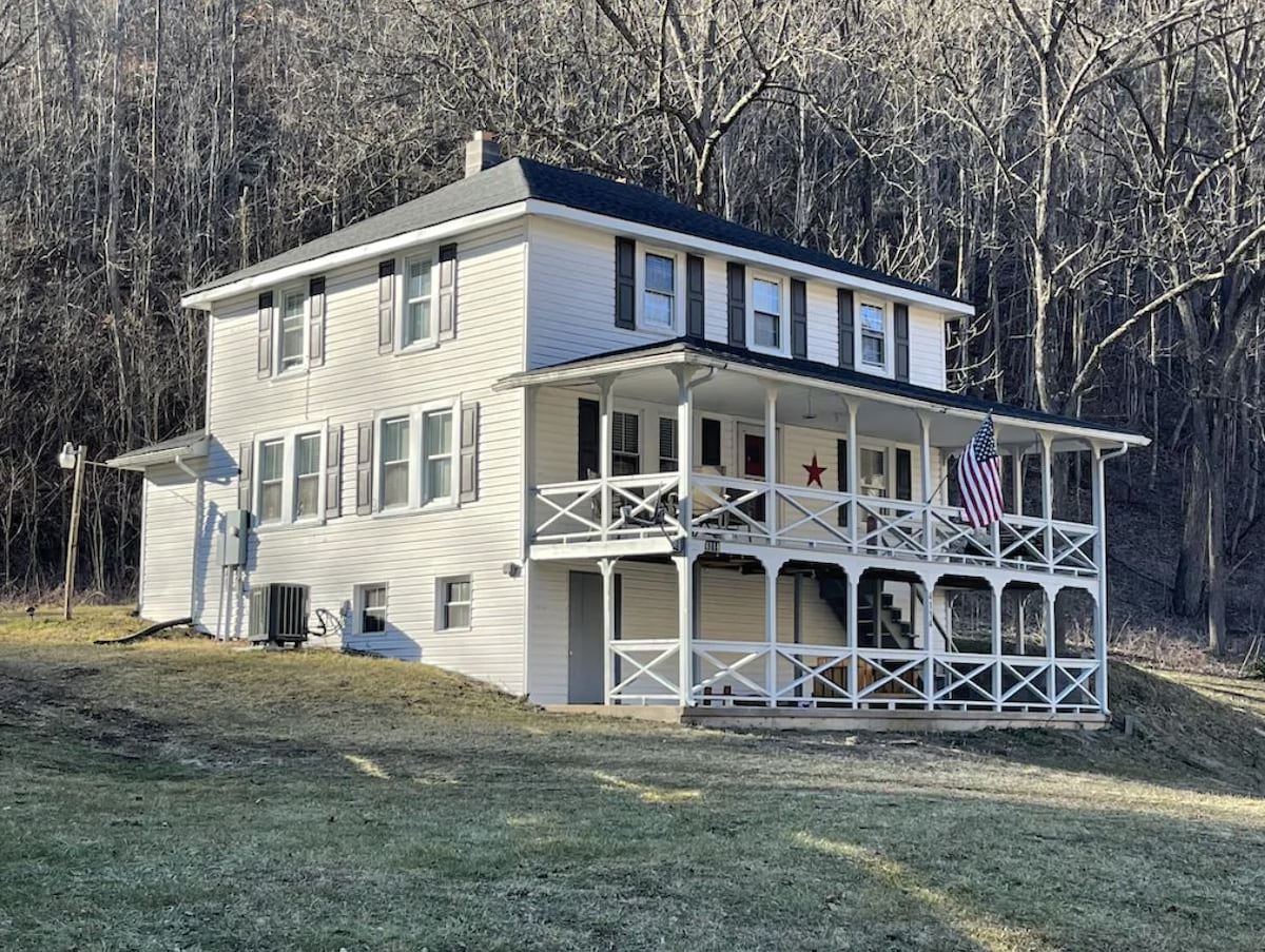 A large, two-story farmhouse is surrounded by a grassy area and wooded landscape. The front porch features white railing and steps leading up to the entrance. An American flag is displayed, and ample windows provide a view of the outdoors.
