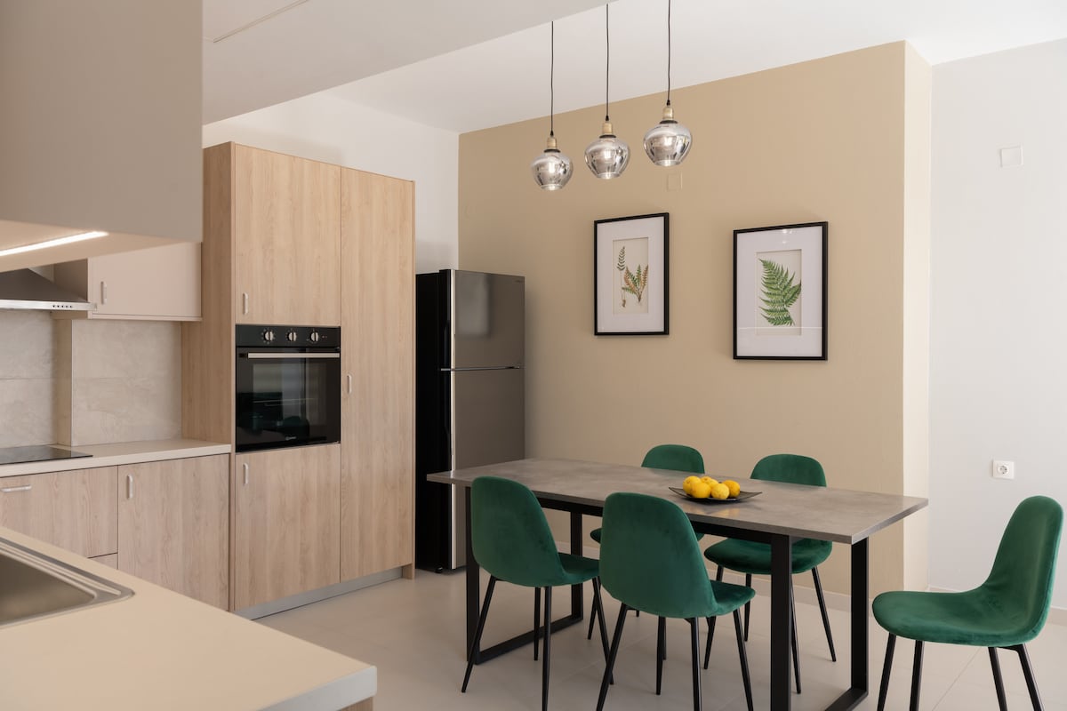 A modern kitchen area features light wood cabinetry and a sleek black refrigerator. A dining table with a stone surface is surrounded by six green chairs. Two framed botanical prints adorn the wall, and warm natural light fills the space.