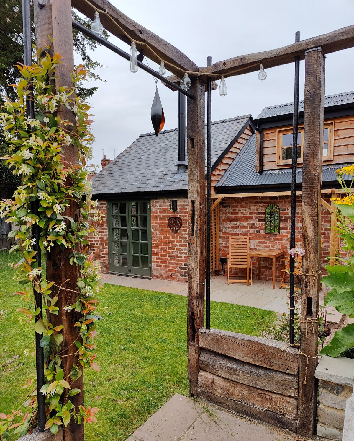 A rustic wooden archway adorned with climbing plants provides an entrance to a quaint garden area. The background reveals a brick cottage with large green doors and a small patio table set for two, inviting outdoor relaxation.
