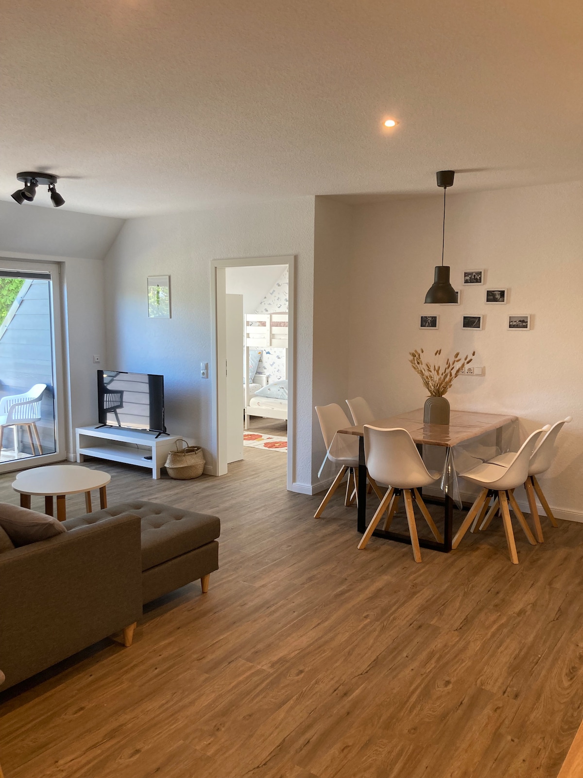 An open living and dining area features a comfortable gray sofa, a glass dining table set for six, and a television on a white stand. Natural light enters through patio doors leading to an outdoor space, enhancing the warmth of the wooden flooring.