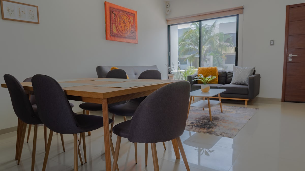 A modern living and dining area features a spacious dining table surrounded by six chairs. A cozy sofa with decorative cushions sits against a large window, offering natural light and views of greenery outside. A small coffee table rests on an area rug.
