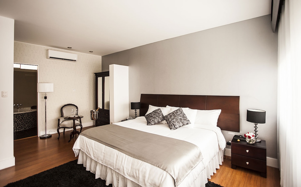 A spacious bedroom features a large bed dressed in crisp white linens, accented by decorative pillows. A lacquered bedside table with a lamp stands next to the bed, while a stylish chair and wardrobe add to the elegant decor. Natural light streams through large windows.