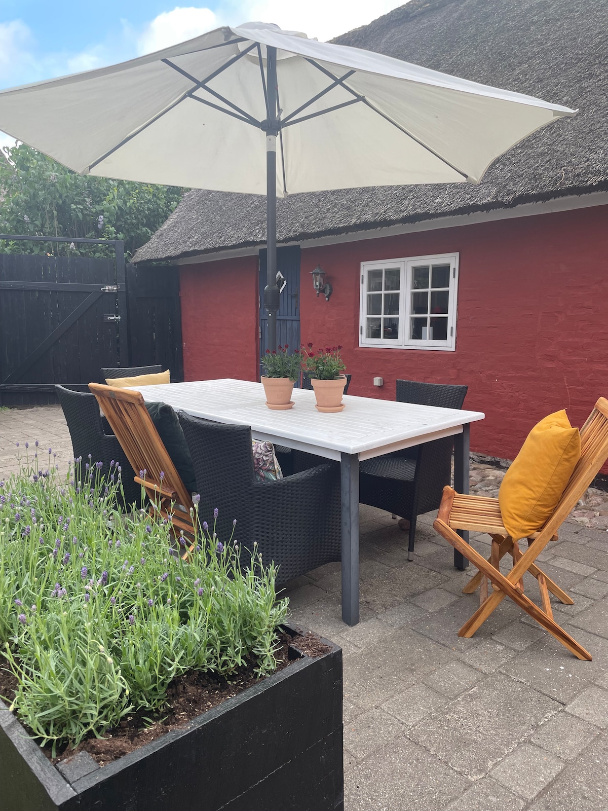 An outdoor dining area features a large table surrounded by six chairs, positioned beneath a large umbrella. Potted plants adorn the table, and a planter with lavender adds a touch of greenery nearby, with a red cottage and wooden fence in the background.