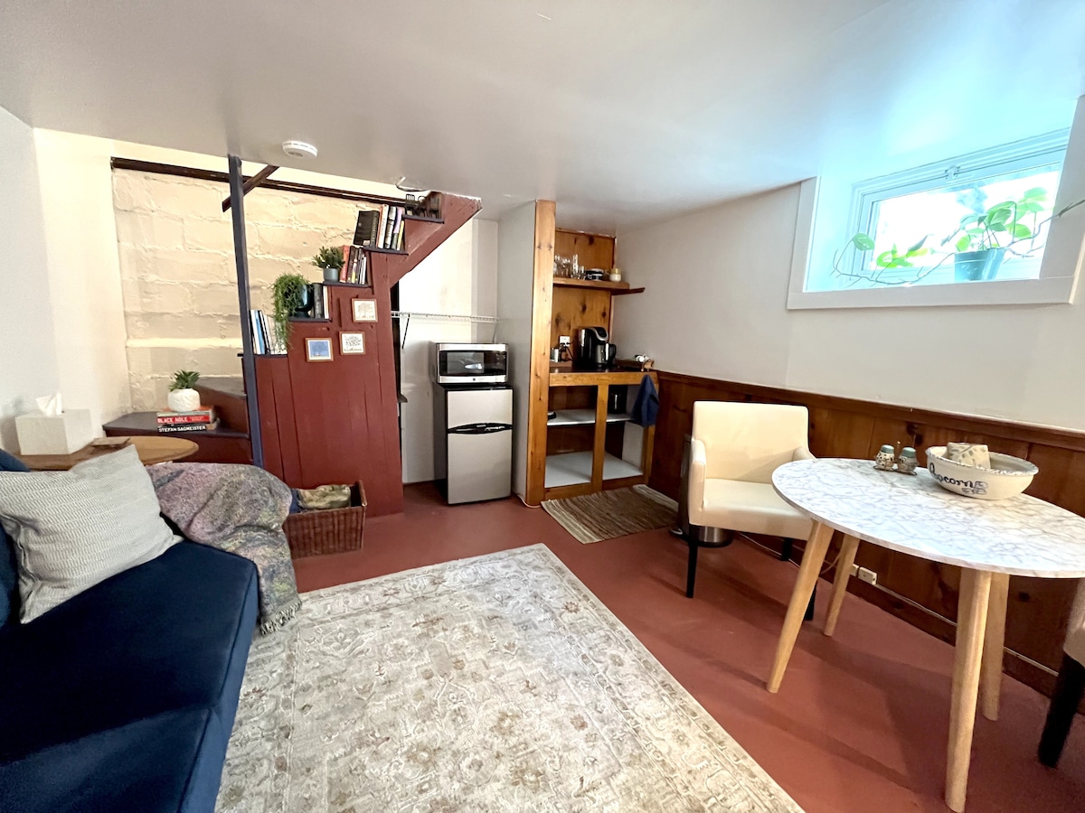 The cozy guest suite features a comfortable couch, a kitchenette with essential appliances, and a dining table. Natural light enters through a small window, highlighting the warm wooden accents and the inviting layout. A plush area rug adds a touch of comfort to the space.