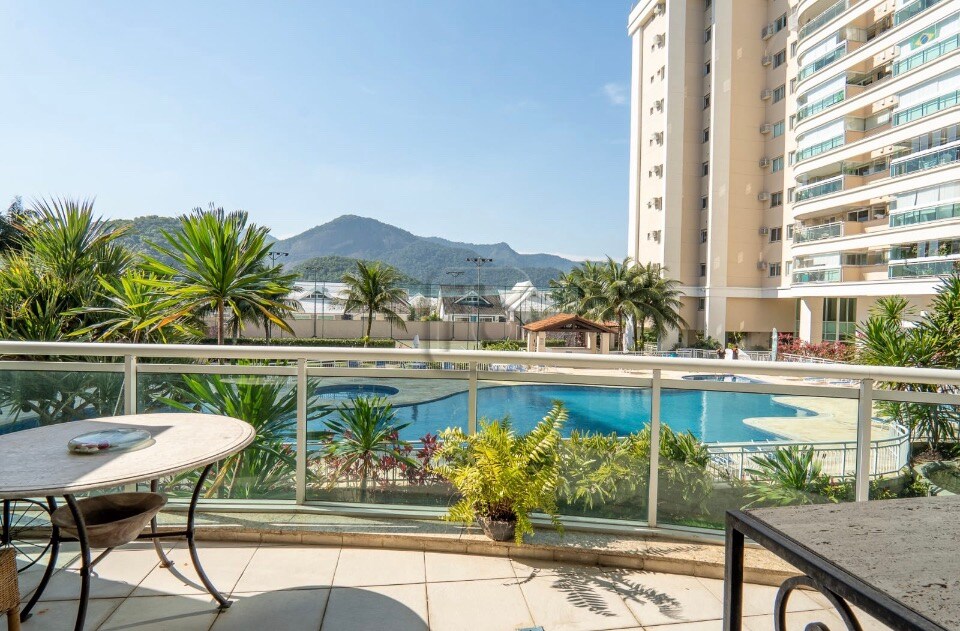 A spacious balcony offers views of lush greenery and a large swimming pool. Outdoor seating is provided with a round table and chairs. The backdrop features distant mountains under a clear blue sky, enhancing the serene atmosphere of the setting.