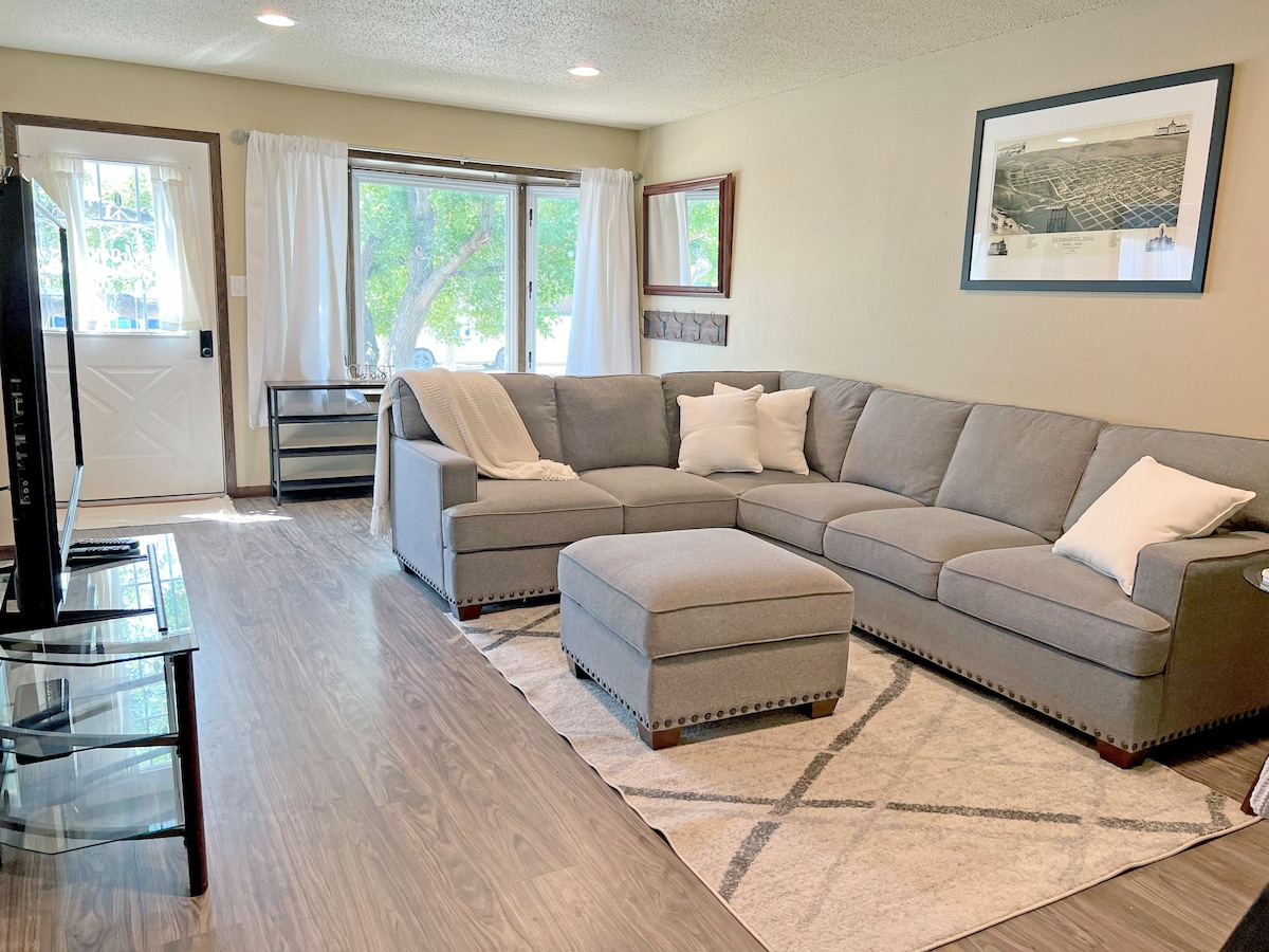 A spacious living area features a large sectional sofa with plush cushions, complemented by a decorative ottoman. Natural light filters through two large windows, and a neutral area rug anchors the seating space. A flat-screen television is positioned on a glass stand across from the seating.