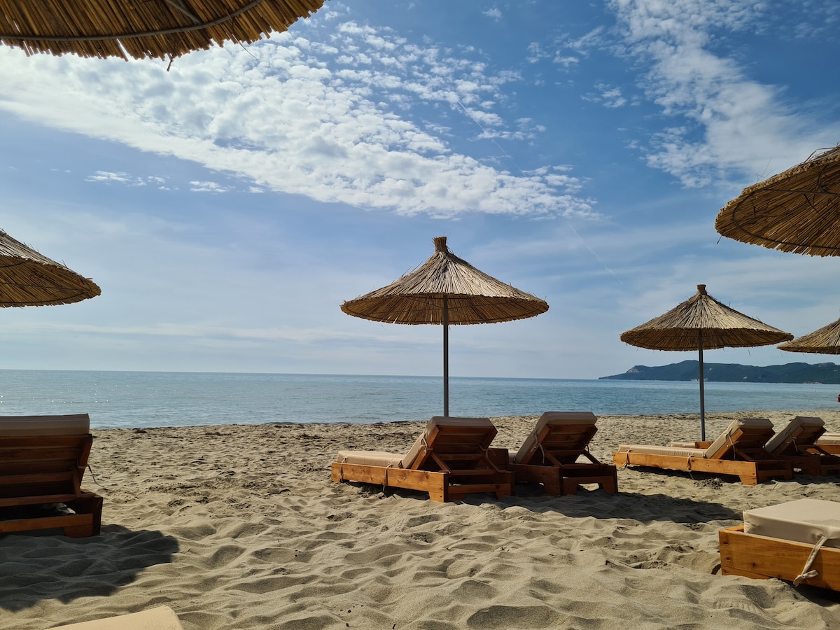 A tranquil beach scene features a row of wooden lounge chairs positioned on fine sand, shaded by large thatched umbrellas. Gentle waves can be seen lapping against the shore in the background, with a clear sky overhead.