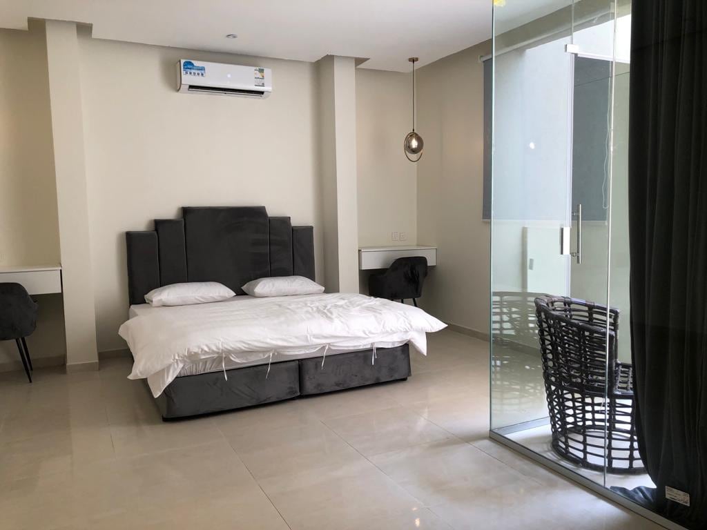 A spacious bedroom features a prominent bed with a light-colored comforter and two white pillows. Natural light enters through a glass door, reflecting on the tiled floor. A modern chair and two minimalist desks are placed against the wall.
