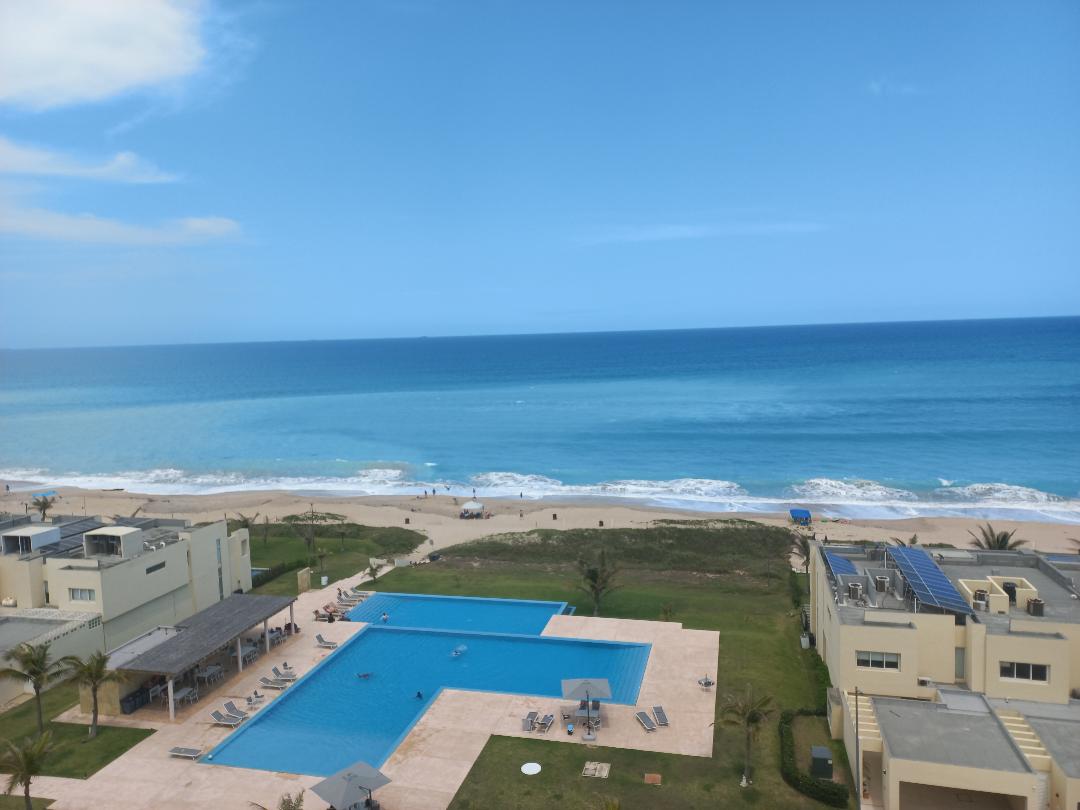 A serene view of the ocean from the sixth floor showcases expansive blue waters meeting the sandy beach. The infinity pool is visible alongside lounge chairs, surrounded by green landscaping. The clear sky adds to the tranquil atmosphere, inviting relaxation by the seaside.