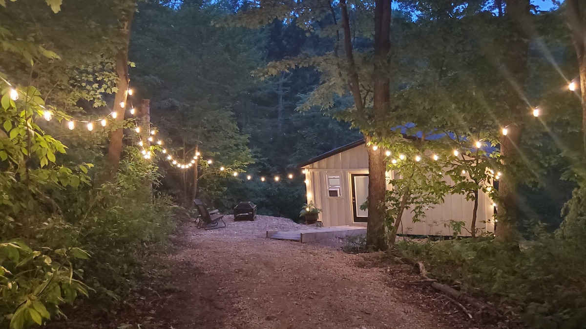 A path lined with glowing string lights leads to a charming lodge nestled among trees. The structure features large windows and a welcoming entrance, surrounded by natural landscaping and gravel. Two outdoor chairs are positioned for relaxation amidst the serene woodland setting.
