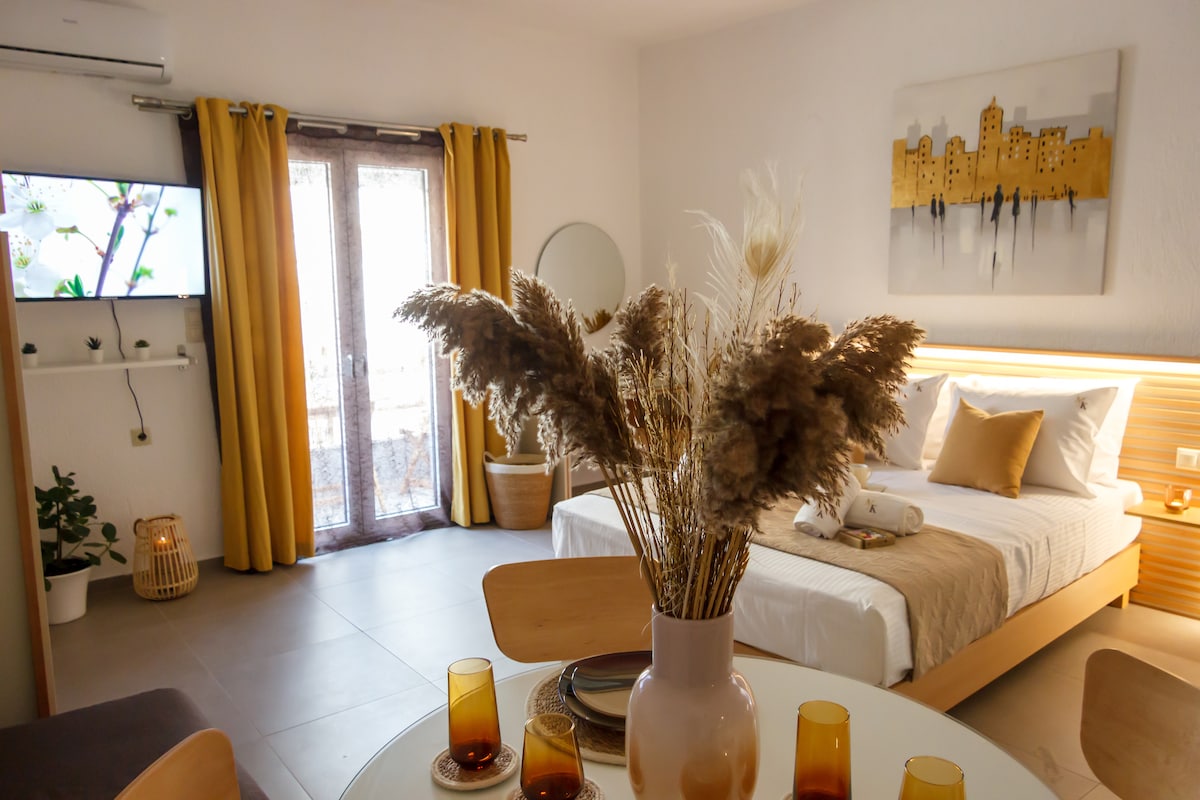 A cozy suite is presented with neutral tones and natural accents. A comfortable bed is situated near a window framed by soft yellow curtains, while a round dining table is adorned with amber glasses and a minimalist vase of dried flowers. A smart TV is mounted on the wall.