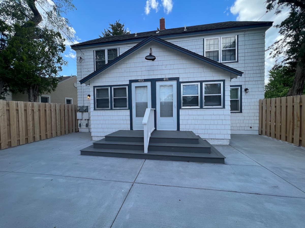 The exterior of the Colonial Duplex is displayed, featuring a white shingled façade with dark trim. A set of steps leads up to the entrance, flanked by large windows. The surrounding area is enclosed by wooden fencing, creating a private outdoor space.