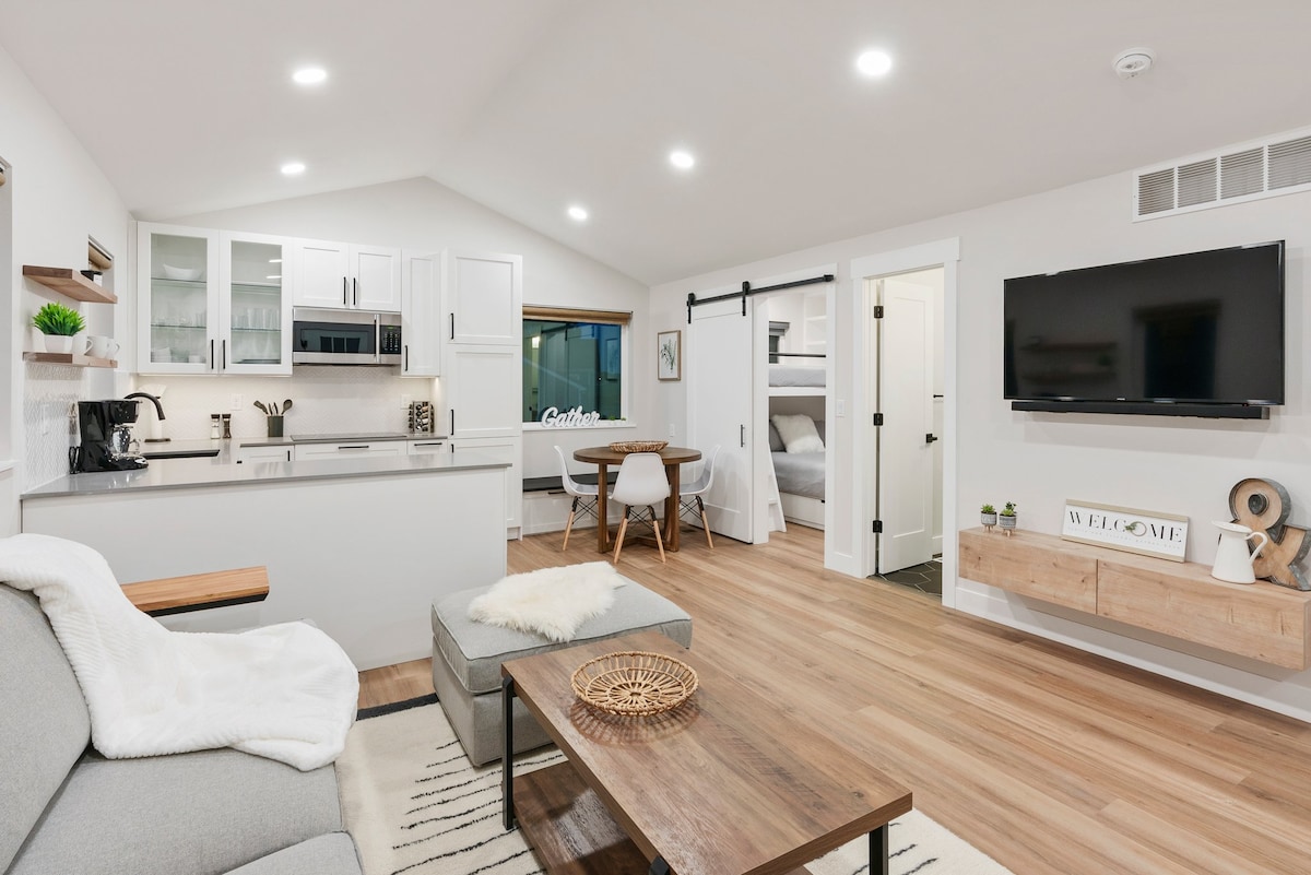 An open-concept living area features a comfortable seating arrangement with a cozy throw. A modern kitchen with light cabinetry is visible in the background, complemented by a dining space with a round table. Sunlight enters through a large window, illuminating the neutral decor.