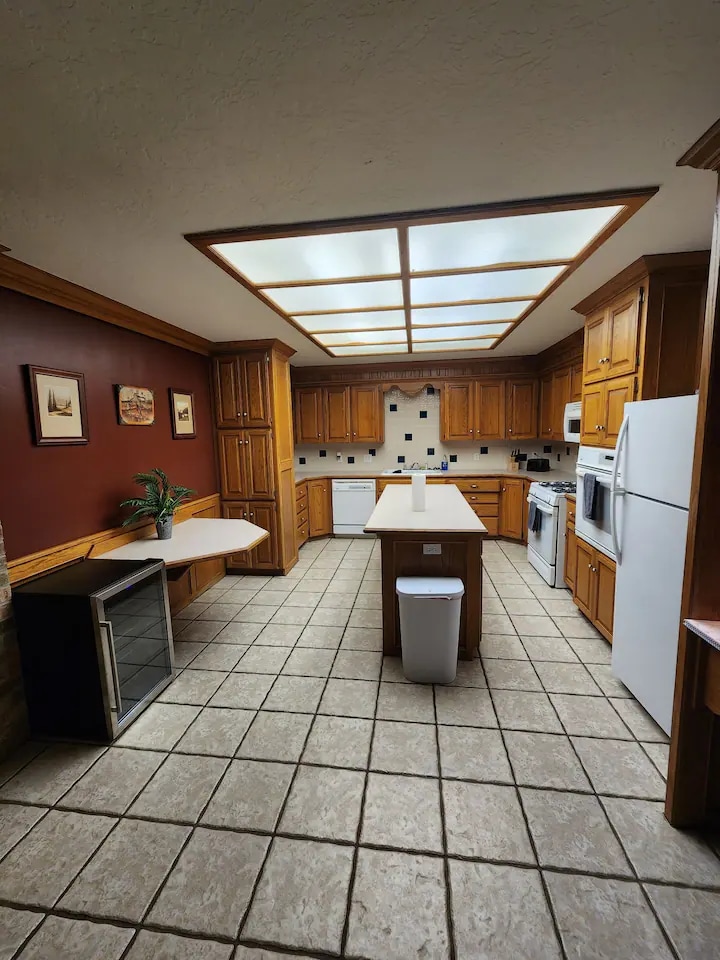 The kitchen features wooden cabinetry and a central island with ample counter space. A set of appliances, including a refrigerator and stovetop, are visible. Natural light filters through a skylight overhead, illuminating the tiled floor and adjacent space for dining or preparation.