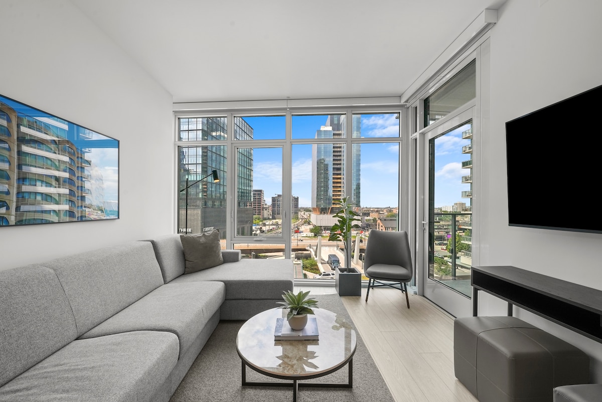 A welcoming living area features a spacious gray sectional sofa and a round coffee table positioned in front of a large window. Natural light floods the space, revealing a view of the city skyline, framed by modern glass architecture.