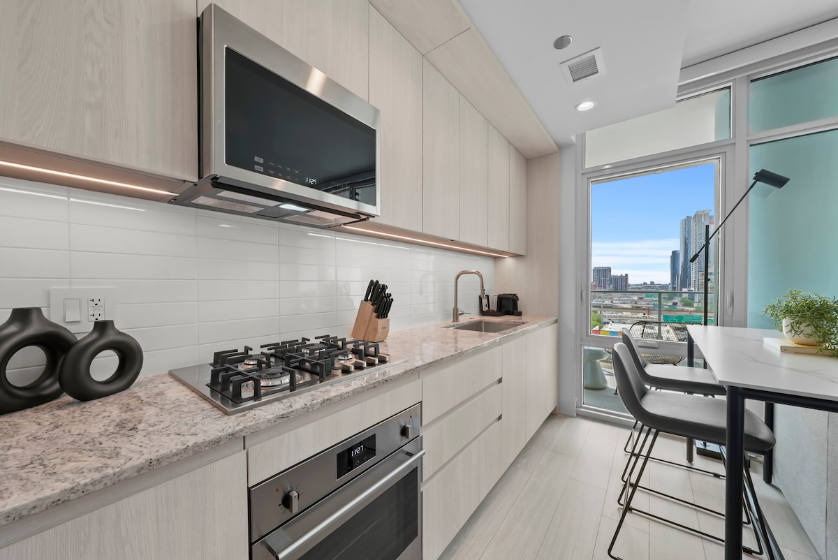 A modern kitchen space is characterized by sleek cabinetry and a light stone countertop. Stainless steel appliances, including a gas cooktop and an oven, are displayed. Natural light floods in through large windows, revealing a vibrant cityscape beyond.