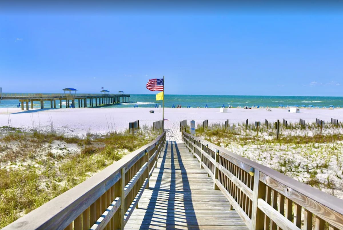 A wooden boardwalk leads directly to the beach, flanked by delicate grasses and sand. An American flag stands proudly at the entrance, overlooking a wide beach area with scattered beachgoers and a distant pier extending into the water.