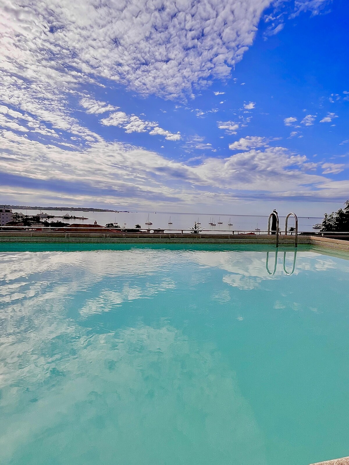The rooftop pool is visible, its surface reflecting the sky and surroundings. The tranquil blue water offers a serene setting with views of the bay and distant boats. The pool area is framed by a comfortable railing, enhancing the peaceful atmosphere.