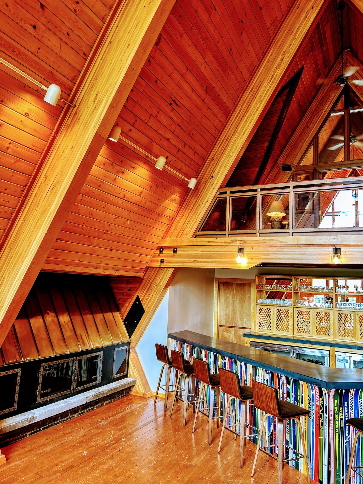 An open-concept lounge area features a high, wooden A-frame ceiling, accentuated by triangular windows. A bar with high stools runs along one side, complemented by vibrant striped decor. A stone fireplace adds a touch of warmth to the space, enhancing the natural wood tones.